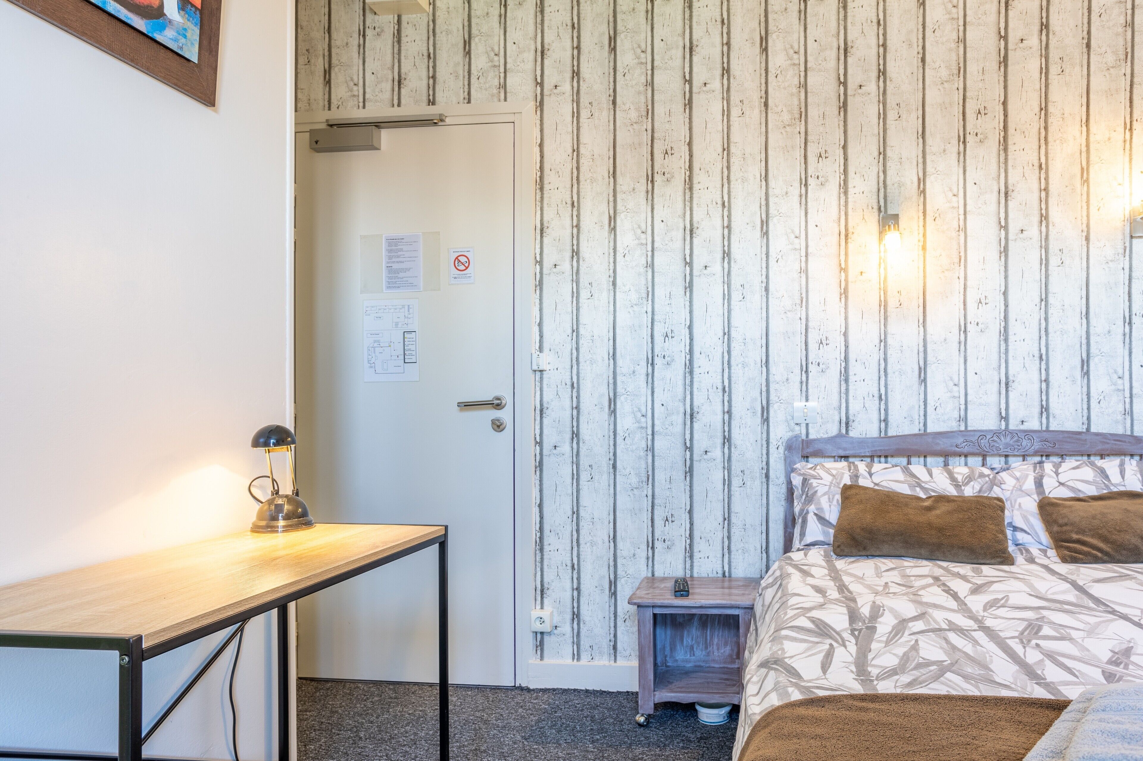 Une pièce comprenant un lit, un bureau, une table de chevet, une porte et un mur orné de lignes verticales.