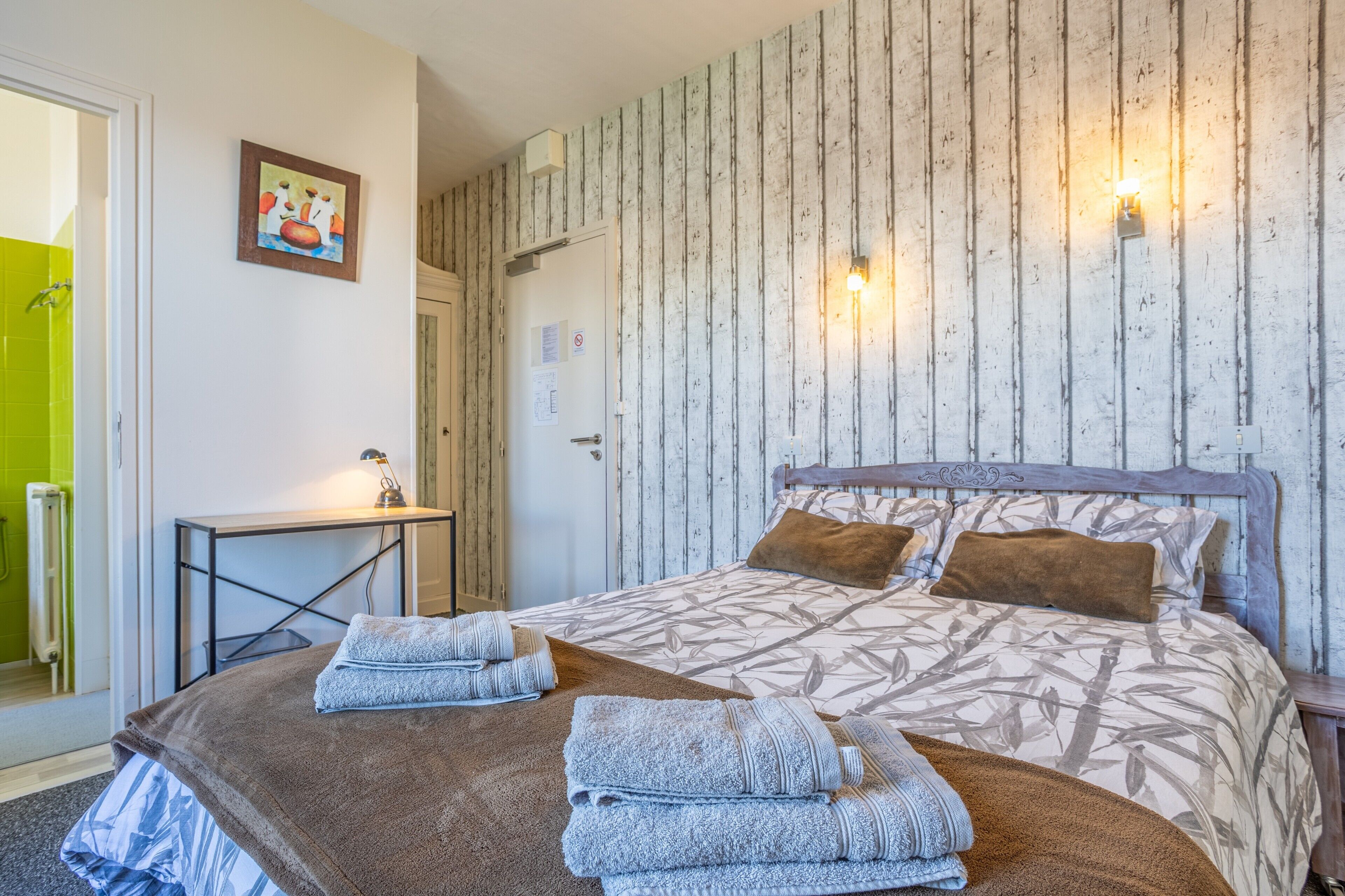 Een slaapkamer met een bed, kussens, een bureau, een stoel, een radiator en een schilderij aan de muur.
