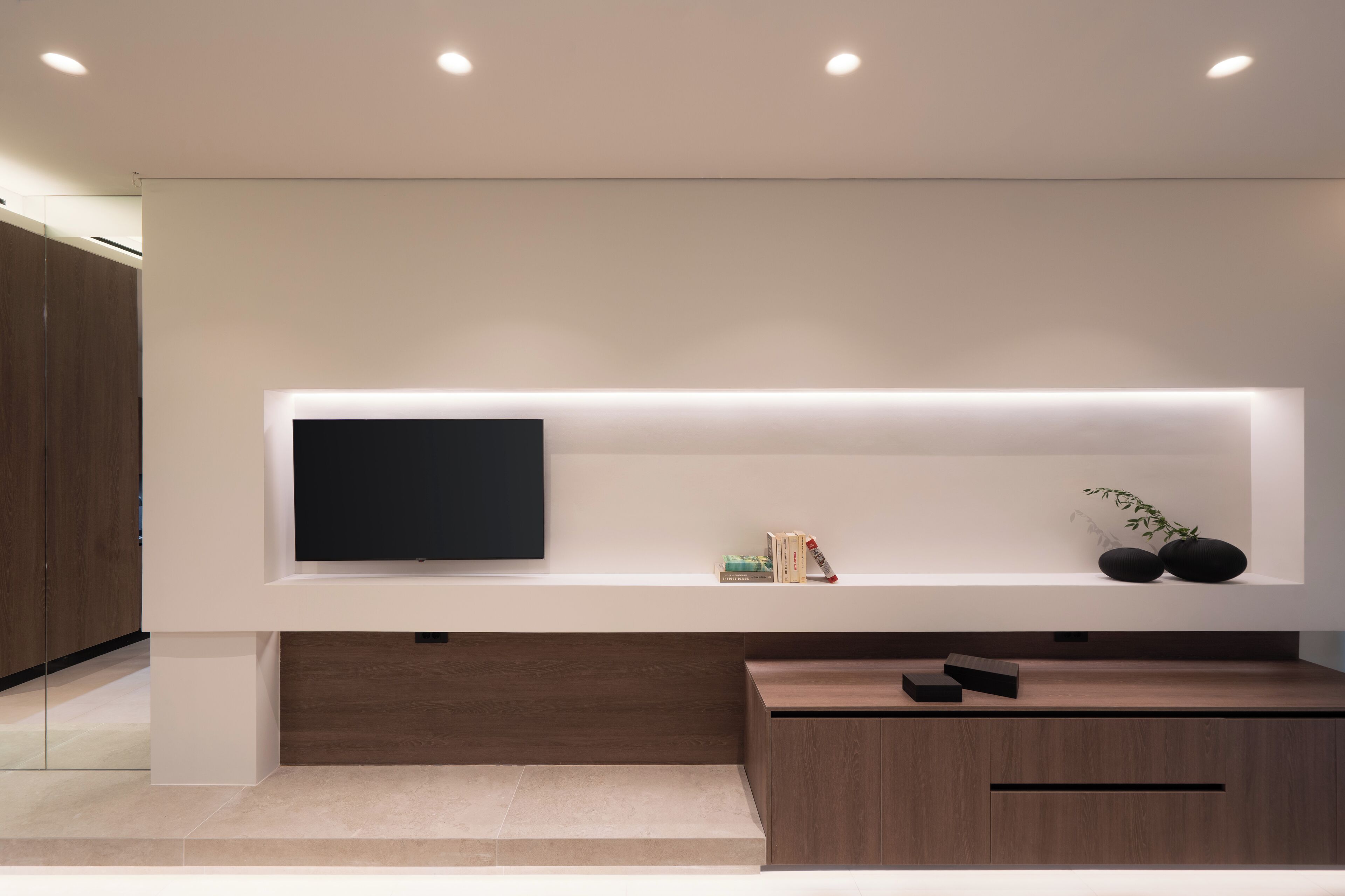 A modern living room with a flat-screen TV mounted on a white wall, a wooden cabinet below, and a small shelf with books and a plant.