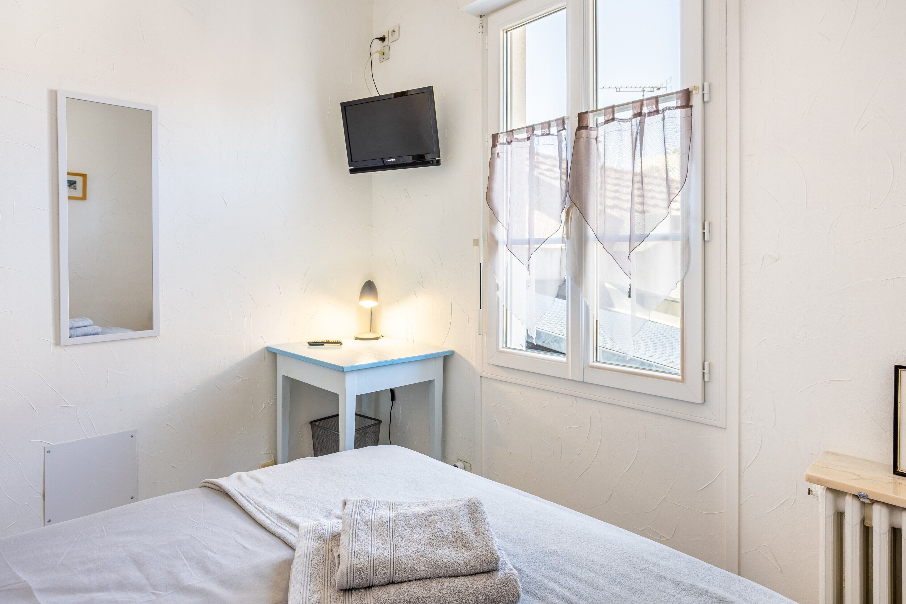 Un dormitorio con cama, escritorio con lámpara, televisor montado en la pared y ventana con cortinas.