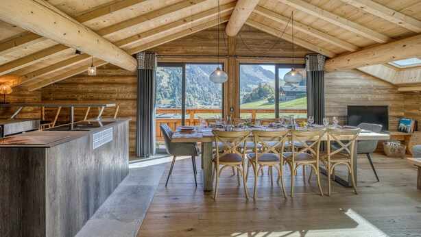A spacious dining area with a high ceiling and wooden beams, a large window overlooking a mountainous landscape, and a television on the right side. - "Borderan" Apartment with Mountain Views, Jacuzzi, Balcony, and Wi-Fi (La Clusaz)