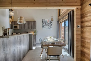 A modern kitchen with a built-in microwave, a dining area with a wooden table set for four, and a large window with curtains. - Apartment "L'Etale" with Mountain Views, Jacuzzi, Balcony, and Wi-Fi (La Clusaz)