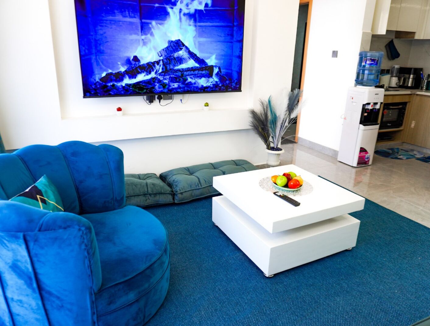 A modern living room with a blue sofa, a white coffee table with fruit, and a TV displaying a fire scene.