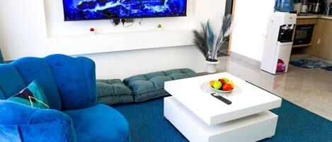 A modern living room with a blue sofa, a white coffee table with fruit, and a TV displaying a fire scene.