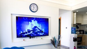 Una sala de estar moderna con un sofá azul, una mesa de centro blanca con fruta, y un televisor montado en la pared mostrando una imagen de una chimenea.