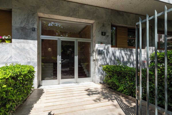 Una entrada de edificio moderno con puerta de vidrio, reja metálica y una escalera que conduce al interior.