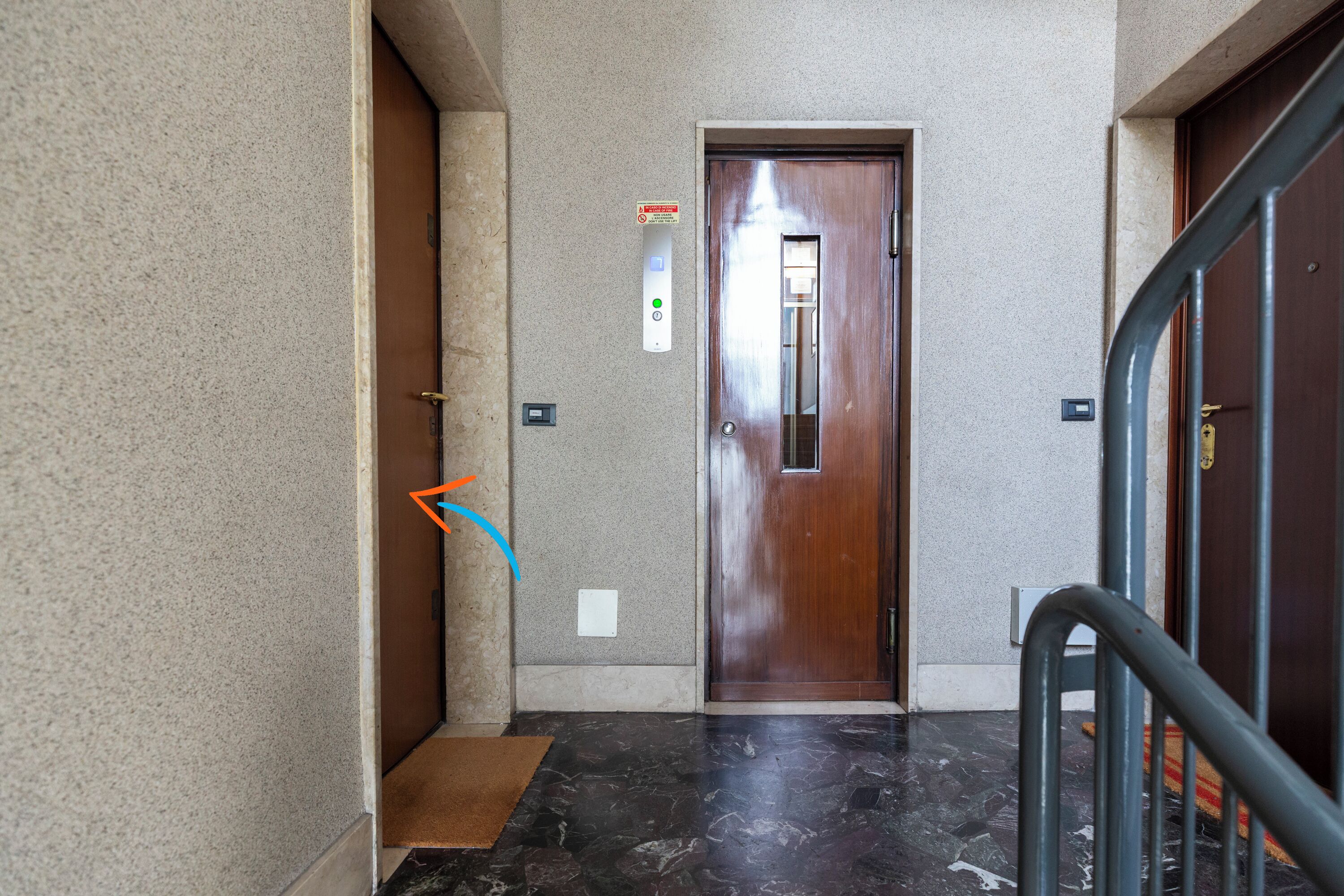 A hallway with a wooden door, a wall-mounted elevator control panel, and a metal handrail.