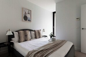 Una cama bien hecha con edredón blanco, cabecera oscura y varias almohadas. Hay una mesita de noche con una lámpara y un pequeño jarrón con flores. Un cuadro enmarcado cuelga de la pared sobre la cama.