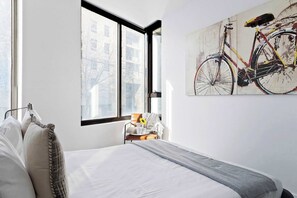 Ein Schlafzimmer mit Bett, einem Sessel mit Tisch, einem Fenster und einem an der Wand hängenden Bild von einem Fahrrad.