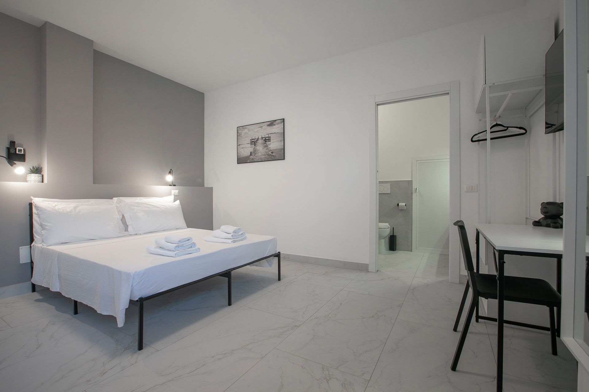 A modern hotel room with a bed, bedside table, and a desk with a chair.