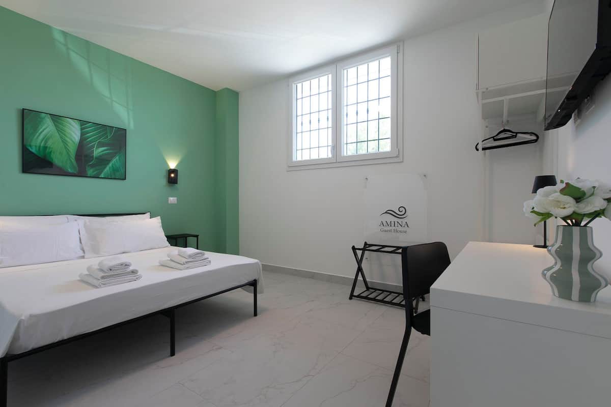 A modern guest room with a bed, a desk, a chair, a window, and a wall-mounted light fixture.