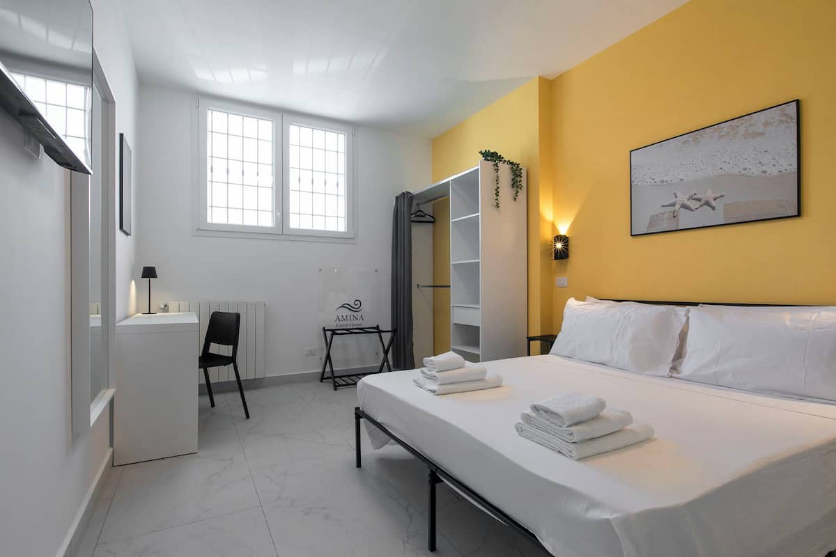 A modern guest room with a bed, bedside table, and a desk with a chair. There is a wardrobe with a mirror, a window, and a wall-mounted light fixture.