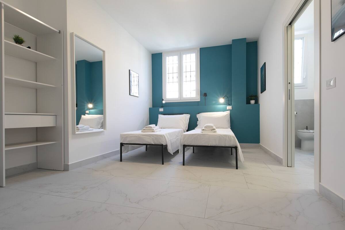 A modern hotel room with two beds, a large mirror, a wardrobe, and a bathroom visible through a glass door.