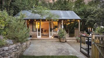 A rustic cabin with a stone wall, a wooden deck, and a cozy interior with a sofa and cushions.