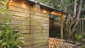 A wooden cabin with a stone wall and wooden deck. There are plants and a wooden bench.