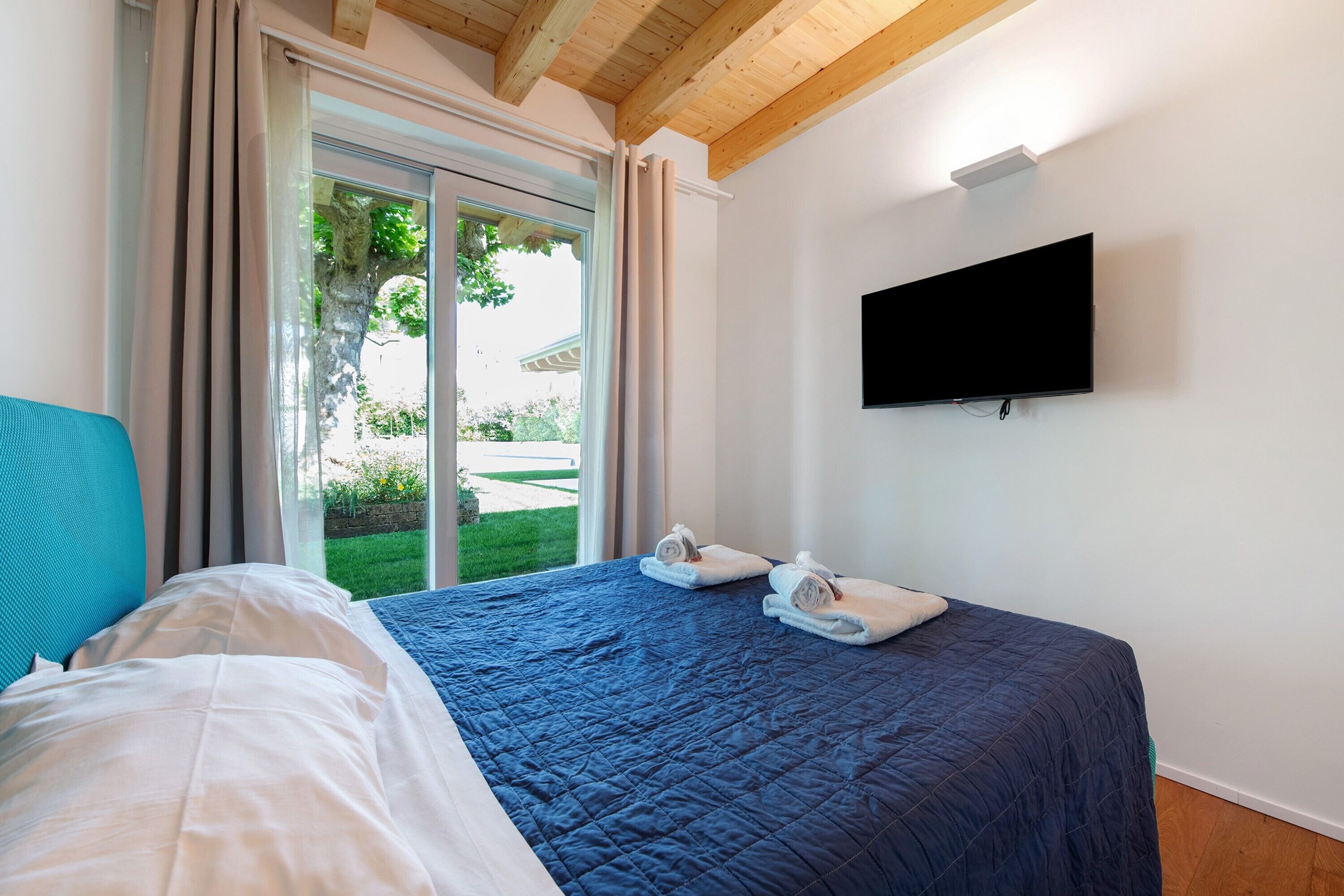 A bedroom with a bed, a TV mounted on the wall, and a sliding glass door leading to a garden.