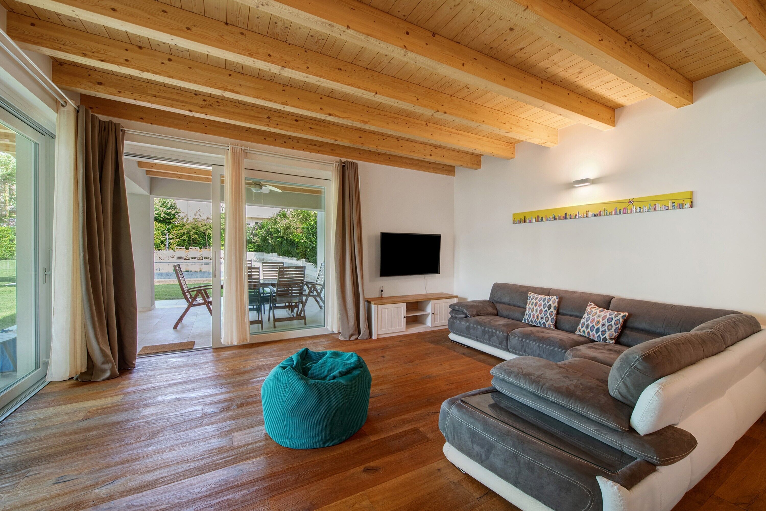 Un salon moderne avec des poutres en bois, un canapé gris, un pouf turquoise et un téléviseur fixé au mur.