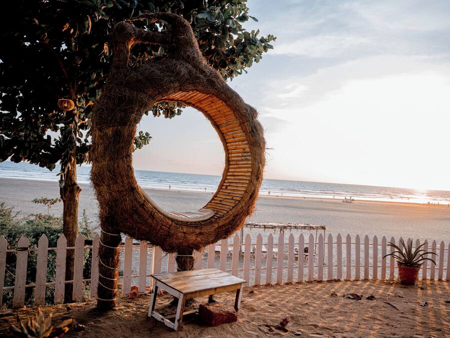 Escena de playa con una estructura circular tipo espejo, cerca hay una valla de piquetes blancos y un banco.