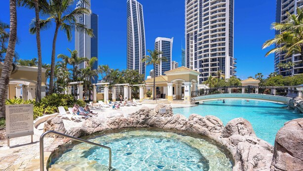 A resort pool with clear water, surrounded by palm trees and modern high-rise buildings in the background.