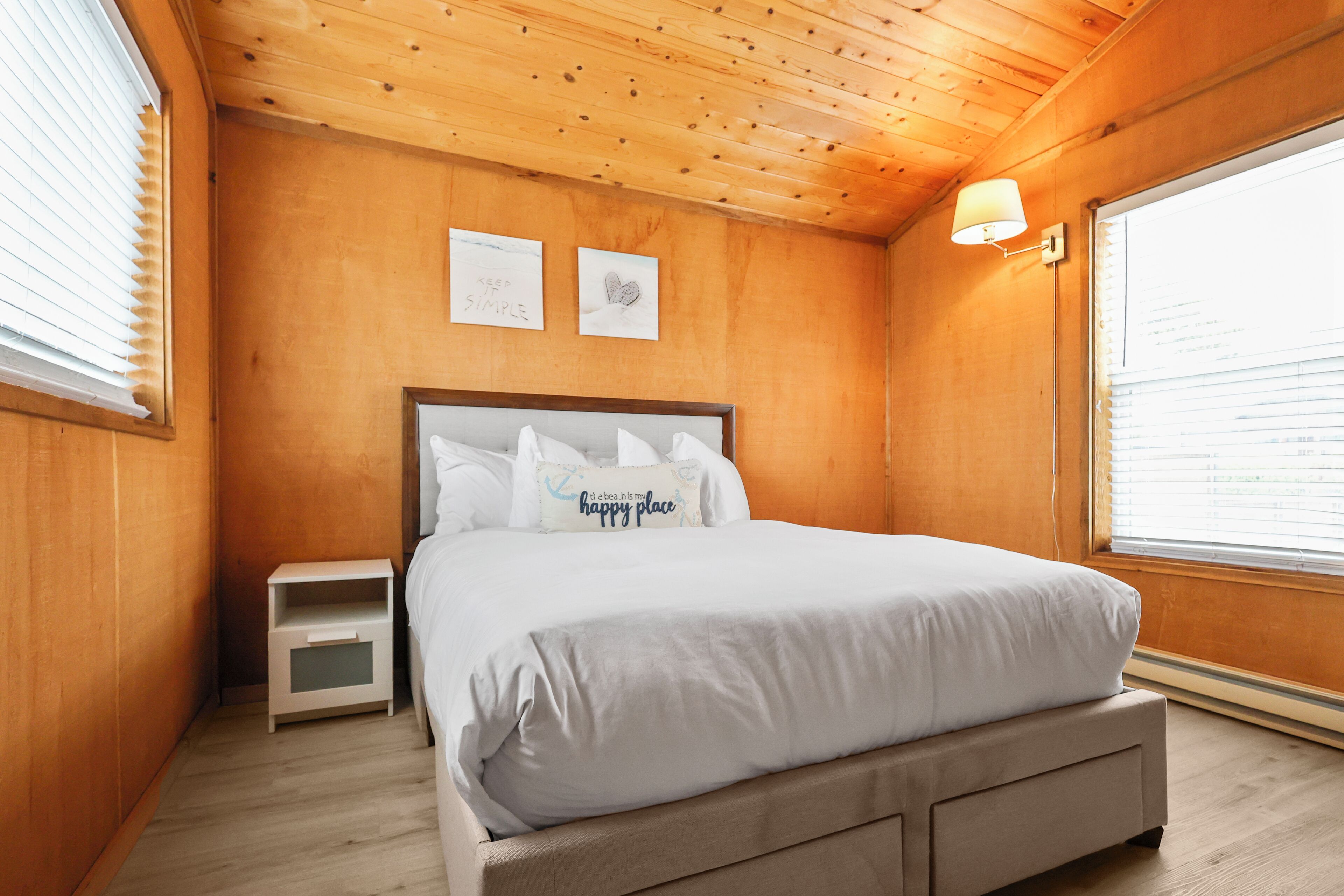 Une chambre aux murs lambrissés, avec un lit, des tables de chevet et une fenêtre équipée de stores.