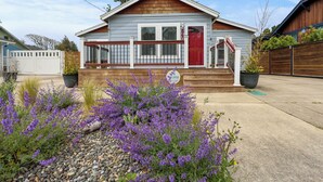 Ein eingeschossiges Haus mit roter Tür, einer kleinen Veranda mit Geländer und einem Garten mit violetten Blumen.