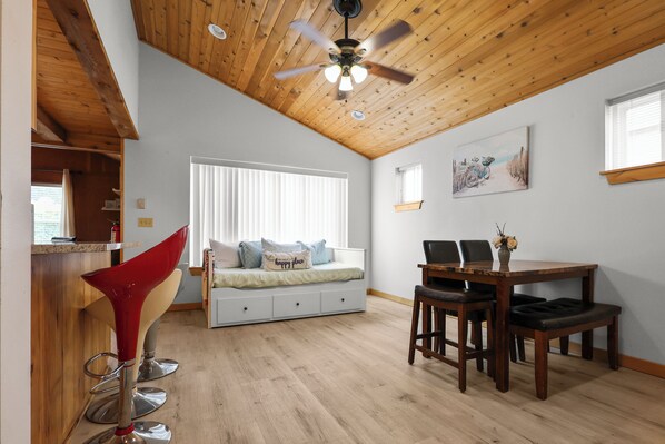 A room with a wooden ceiling, a bed with a window, a dining table with chairs, and a red chair. - 2 Blocks to Beach • Fenced Yard • Pet Friendly (Seaside)