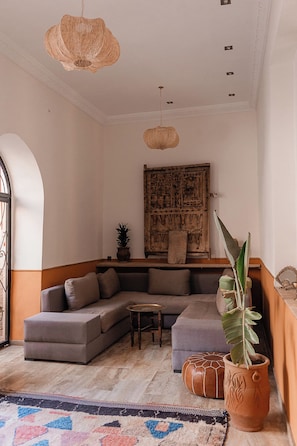 A living room with a sectional sofa, a coffee table, a patterned rug, a potted plant, and a decorative wall hanging. - Le Riad Medina Privilège & Spa (Marrakech)