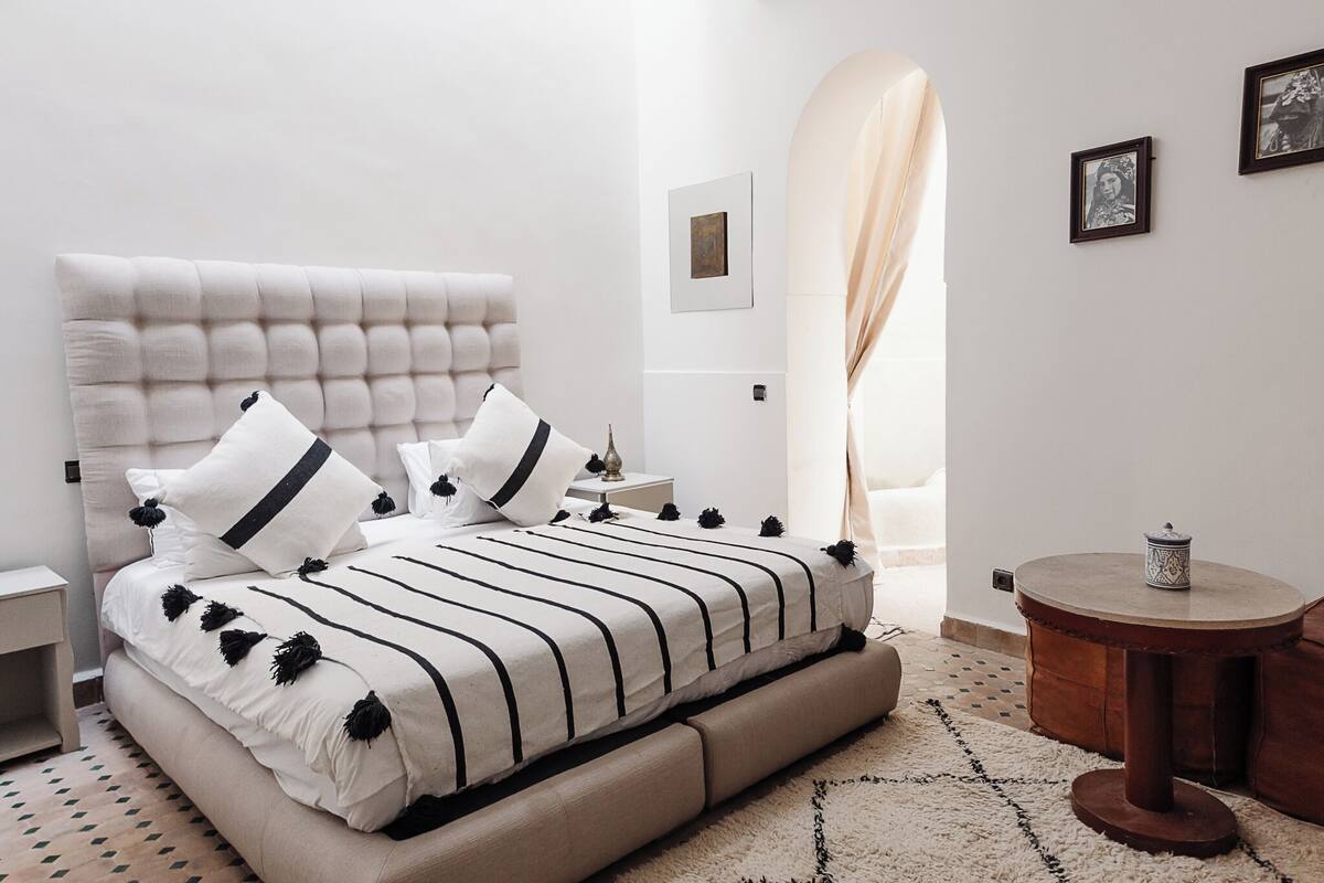 A modern bedroom with a tufted headboard, a large bed with black and white striped bedding, a round wooden side table, and a small round table with a decorative item.
