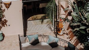 A curved sofa with cushions, a round coffee table, and a potted plant in a courtyard with a patterned floor and a red wall. - Le Riad Medina Privilège & Spa (Marrakech)