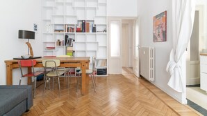 Una habitación con piso de madera, una mesa de comedor con sillas, una estantería, un radiador y una puerta que da a otra habitación.