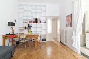 A room with a wooden floor, a dining table with chairs, a bookshelf, a radiator, and a door leading to another room.