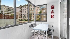 A corner room with a white table and chairs, large windows with a view of apartment buildings, and a 'BAR' sign on the wall. - Mural Oasis w Sunroom, Parking, near Stampede (Calgary)