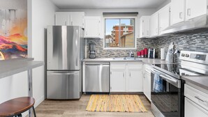 A modern kitchen with stainless steel appliances, white cabinets, a window with a city view, and a colorful rug. - Mural Oasis w Sunroom, Parking, near Stampede (Calgary)