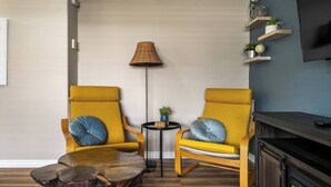 Una sala moderna con dos sillones amarillos, una mesa redonda de centro, una lámpara de pie, una pequeña mesita auxiliar con una planta y un televisor montado en la pared.