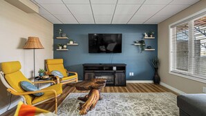 Una sala de estar con un televisor montado en la pared, un sillón amarillo, una mesa de centro de madera y un tapete estampado.
