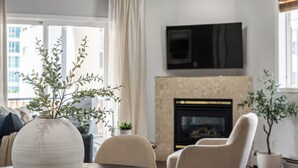 Un salon avec une table ronde en bois, des fauteuils beiges, un vase blanc contenant des plantes vertes, un téléviseur à écran plat fixé au mur et une cheminée.