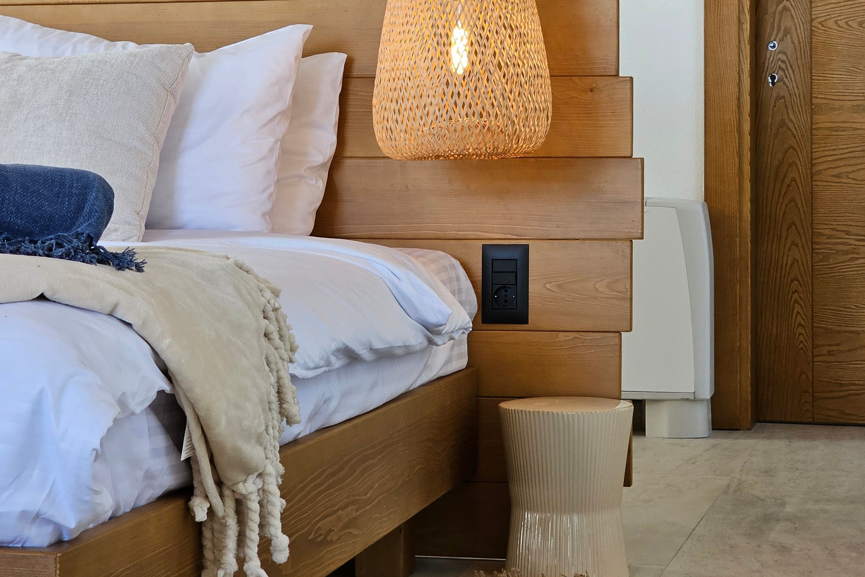 Quarto com cabeceira de madeira, luminária de cabeceira, tomada e um pequeno banquinho.