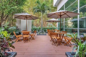 Une terrasse extérieure avec des tables et des chaises en bois, entourée d’une végétation luxuriante et de grands parasols.