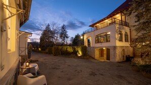 A stone courtyard with outdoor seating, a two-story building with a balcony, and a view of trees and hills in the background.