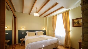 A bedroom with a large bed, wooden beams on the ceiling, a headboard, bedside lamps, a stone wall, and a window with curtains.