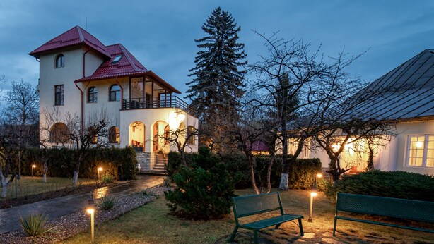 A two-story house with a red roof, a balcony, and a well-lit garden area with benches.