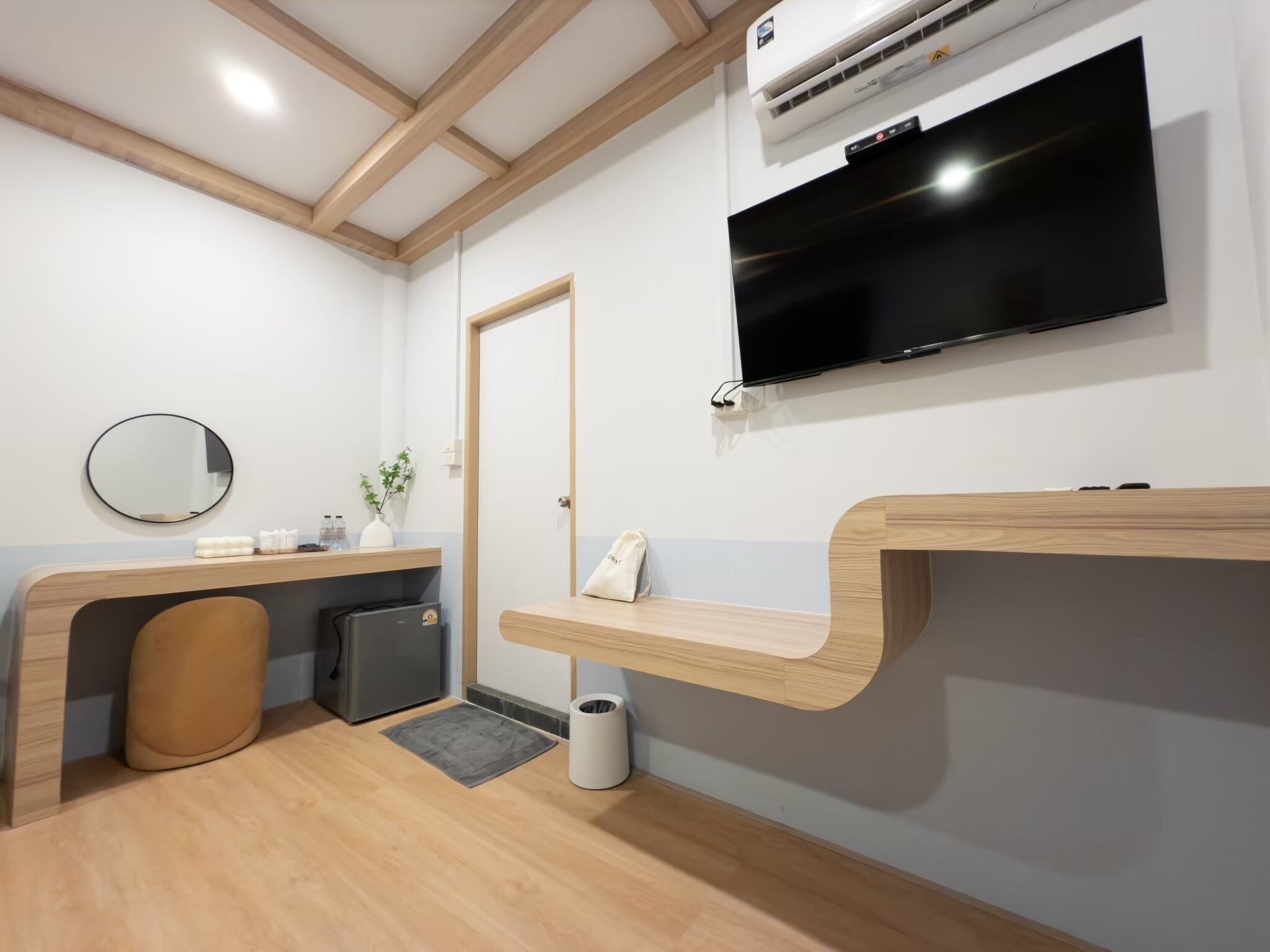 Ein modernes Hotelzimmer mit Holzbalkendecke, einem flachbildfernseher an der Wand, einem runden Spiegel, einem kleinen Schreibtisch mit Blumenvase und Pflegeprodukten sowie einem Mülleimer.