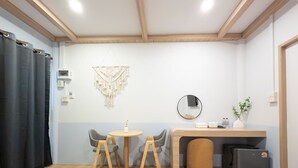 Una stanza piccola e luminosa con soffitto in legno, un tavolo rotondo con due sedie, una scrivania con uno specchio, un frigorifero compatto e un arazzo macramè appeso al muro.