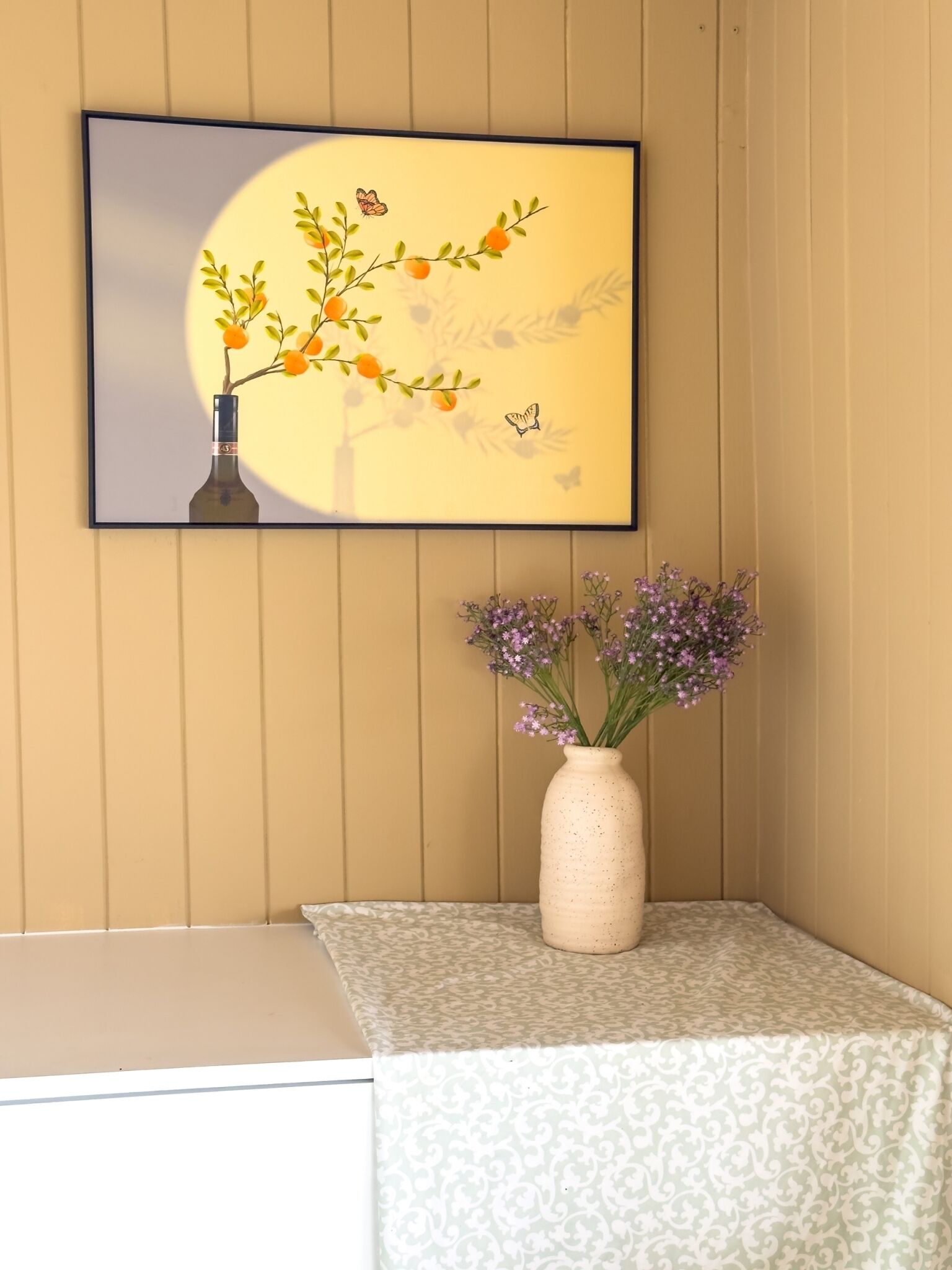 A framed artwork of a tree with oranges and a bottle, a vase with purple flowers, and a patterned tablecloth on a corner table.