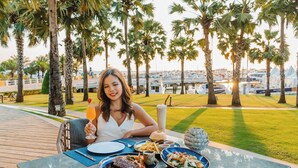 A dining table with food and drinks, palm trees, and a marina in the background.