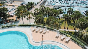 Una gran piscina con sillas de descanso y palmeras, rodeada por un puerto deportivo con barcos.