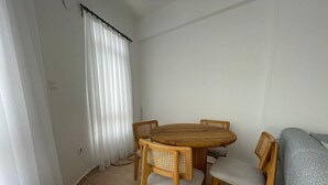 Una habitación pequeña y luminosa con una mesa redonda de madera y cuatro sillas, un sofá gris y una ventana con cortinas blancas.