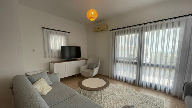 Un salón moderno con un sofá gris, una pequeña mesa redonda para café, un televisor sobre un mueble blanco, y un balcón con cortinas de encaje.
