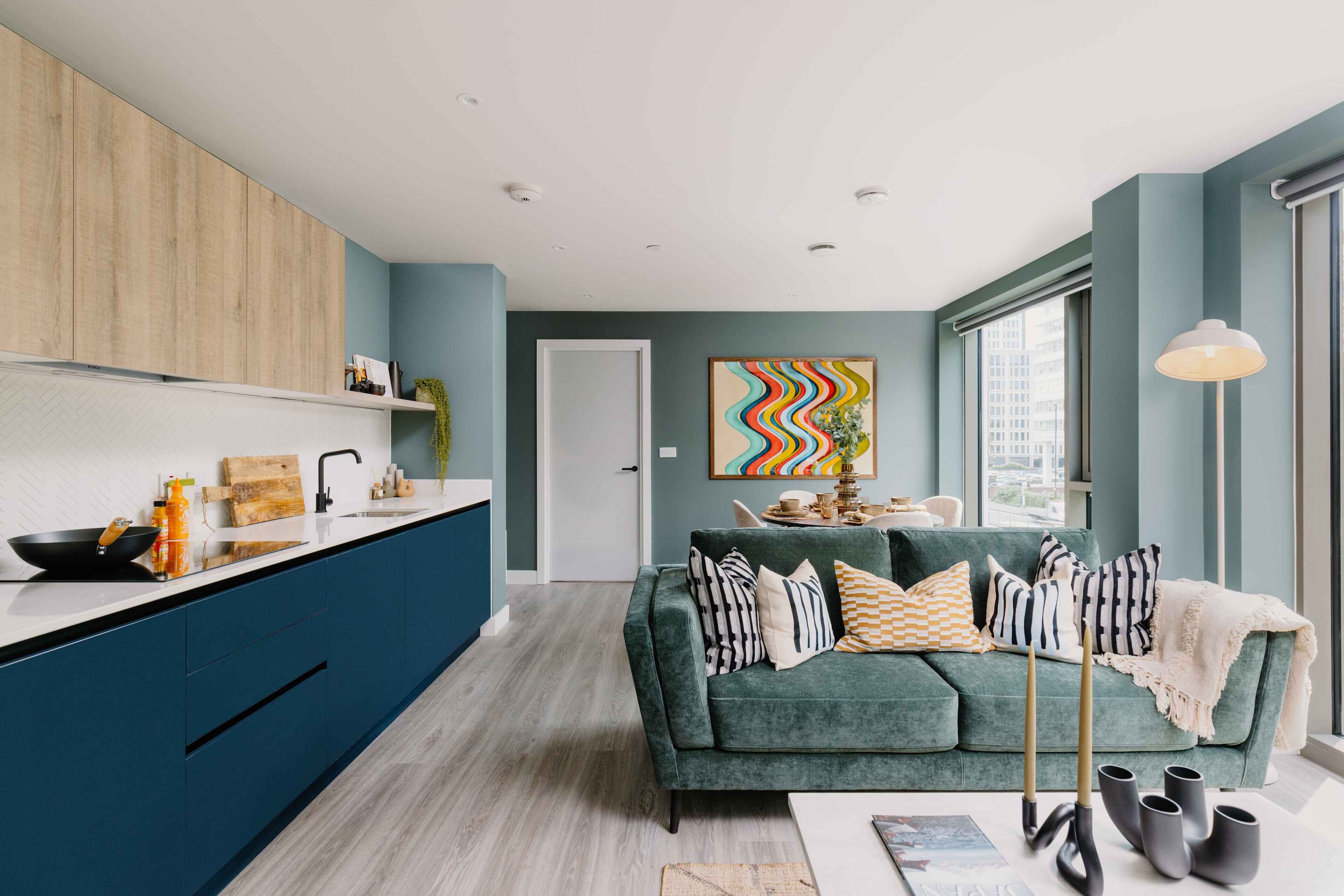 A modern kitchen with blue cabinets, a white countertop, and a sink. There is a green sofa with striped cushions in the living area, and a colorful abstract painting on the wall.
