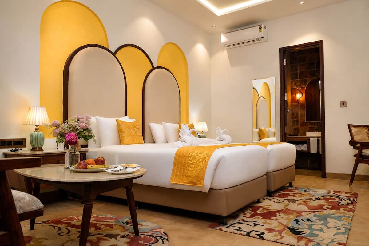 A hotel room with a large bed, a round table with a lamp, a chair, and a bathroom visible through an arched doorway.